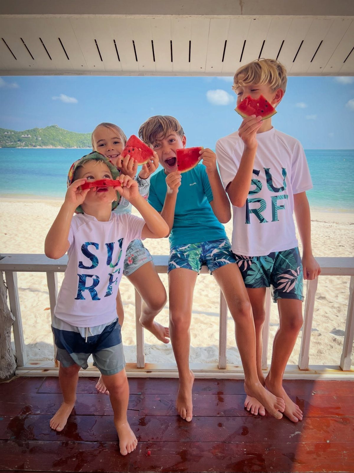Four boys eating watermelon on a deck overlooking a tropical sandy beach