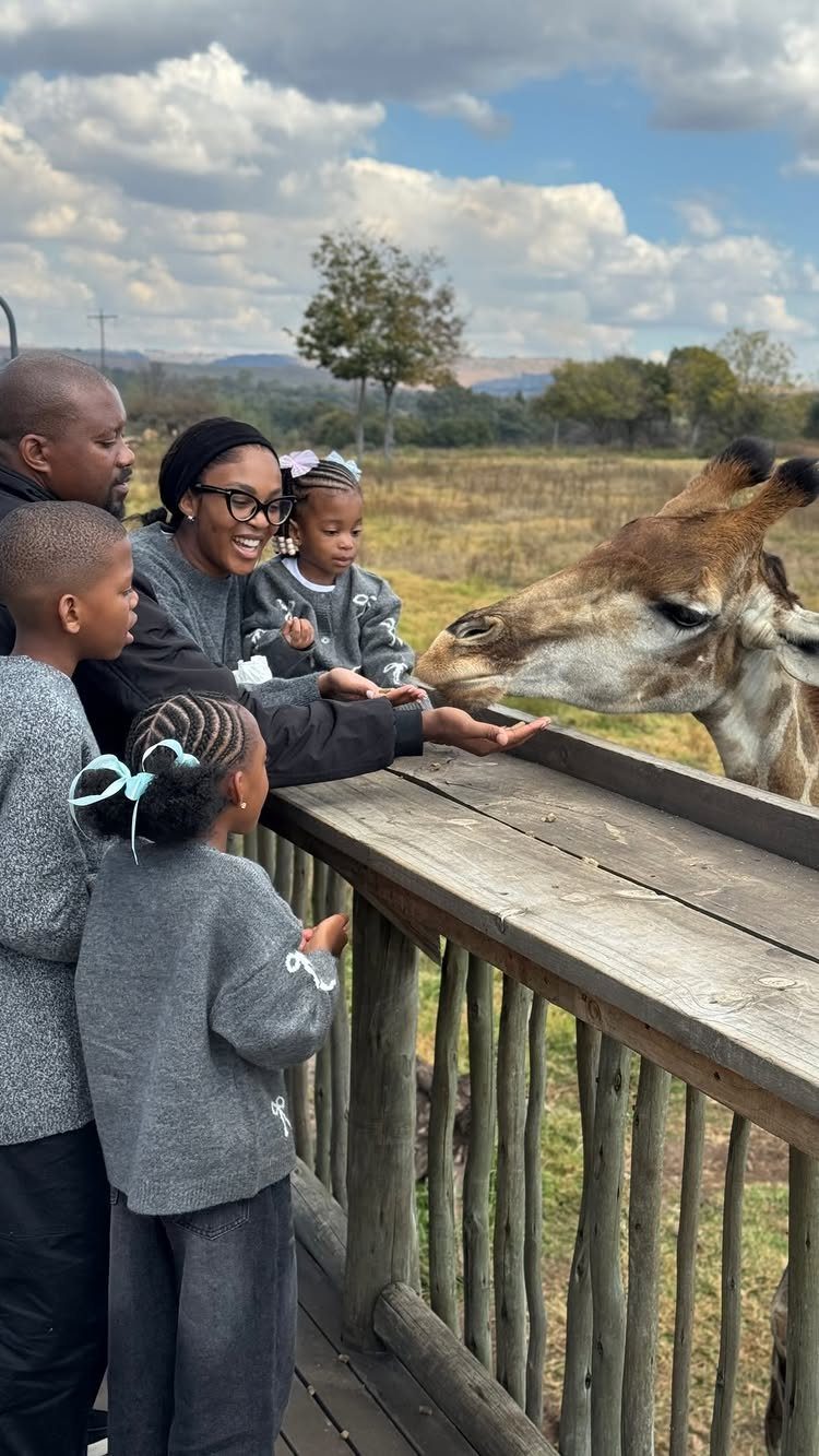 Family feeding a giraffe from a wooden platform at an outdoor animal park.