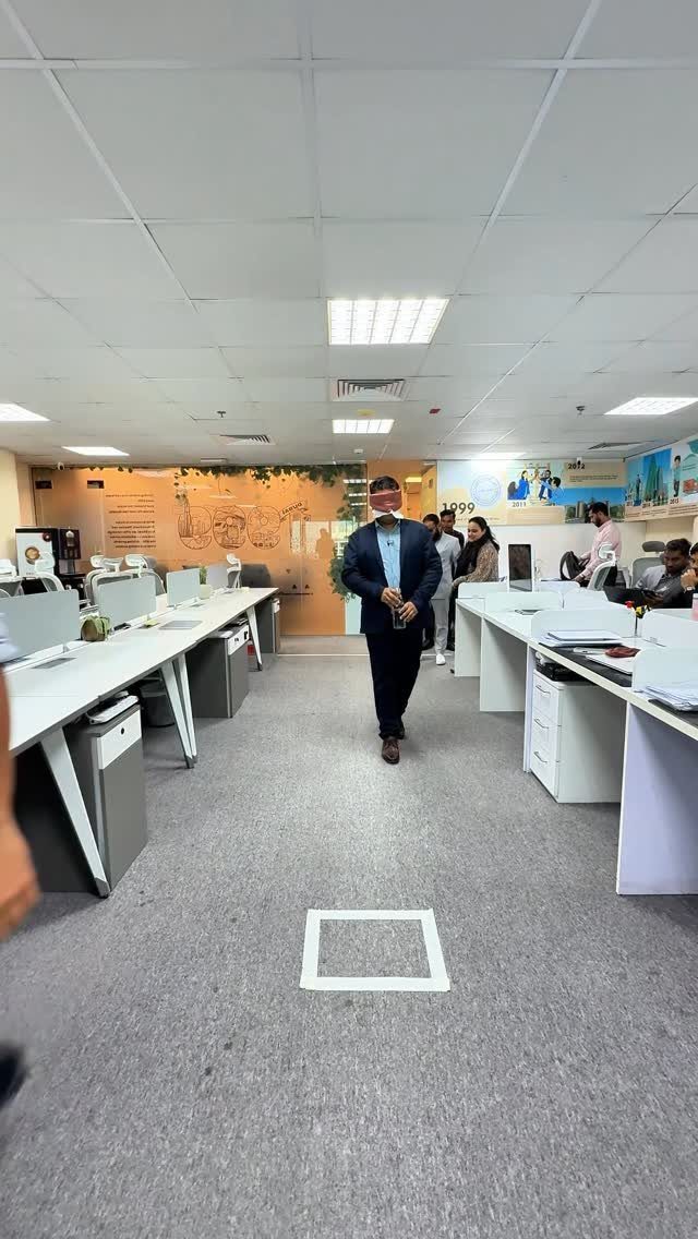 Blindfolded man standing in an office aisle surrounded by white desks and colleagues.