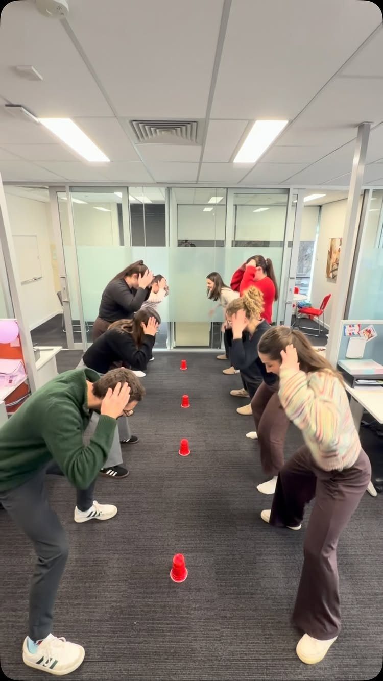 People playing a cup game in an office, bending over with hands on heads.