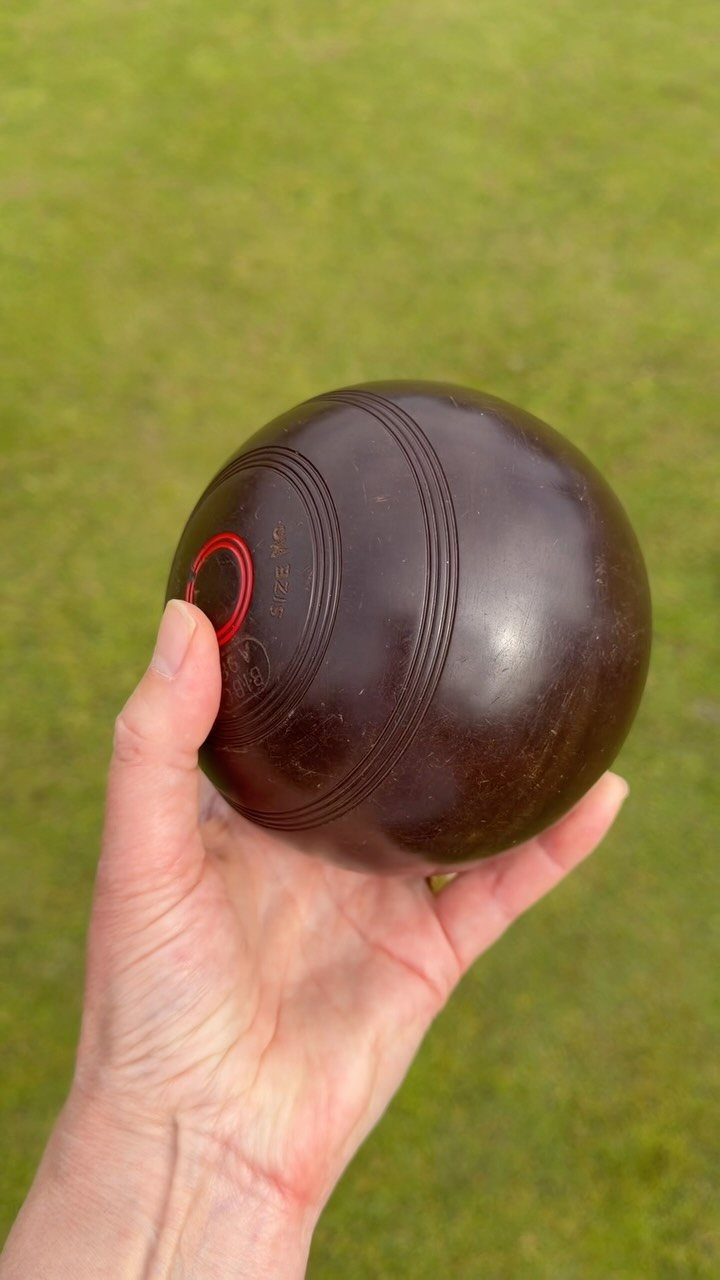 Hand holding a brown bocce ball over a green grass lawn.