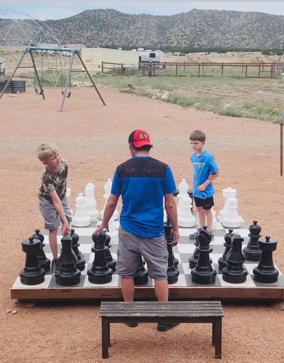 Man and two boys playing on a giant outdoor chessboard in a desert landscape