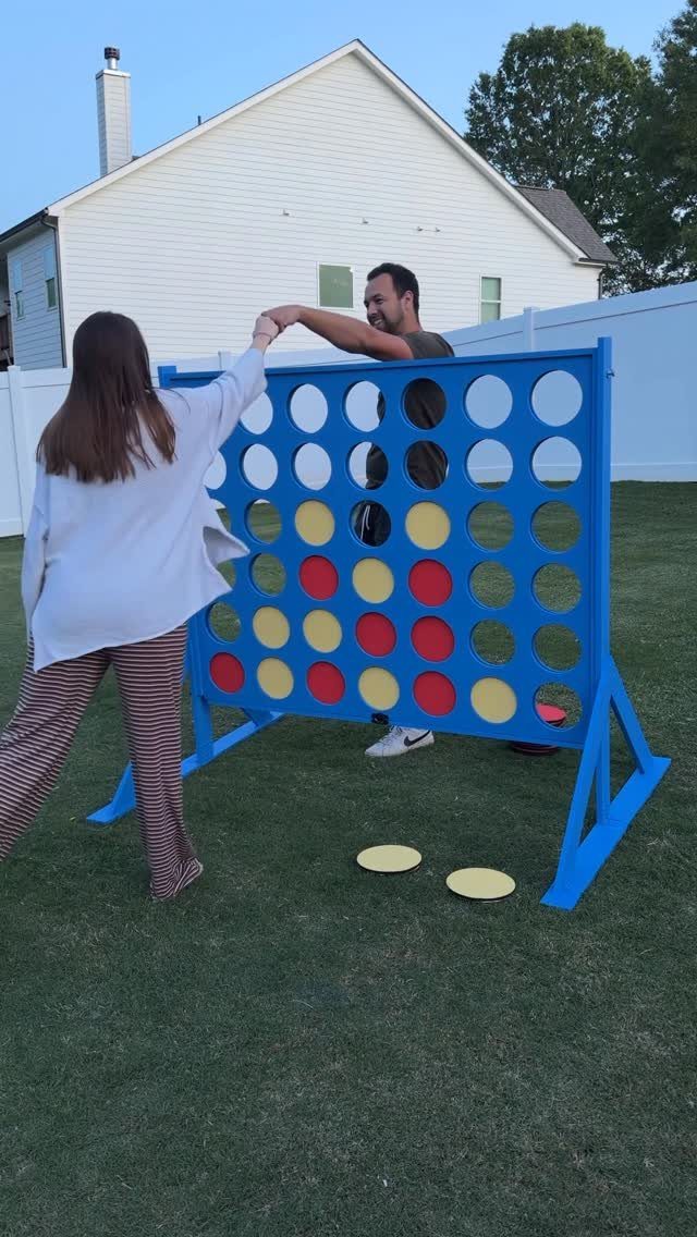 Man and woman standing by a giant blue Connect Four game on a lawn