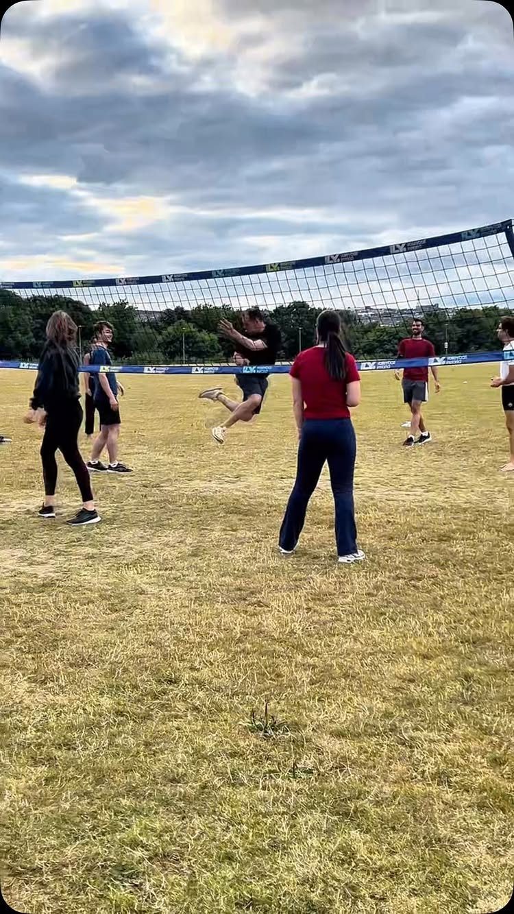 People playing volleyball on a grassy field with a net under cloudy skies.