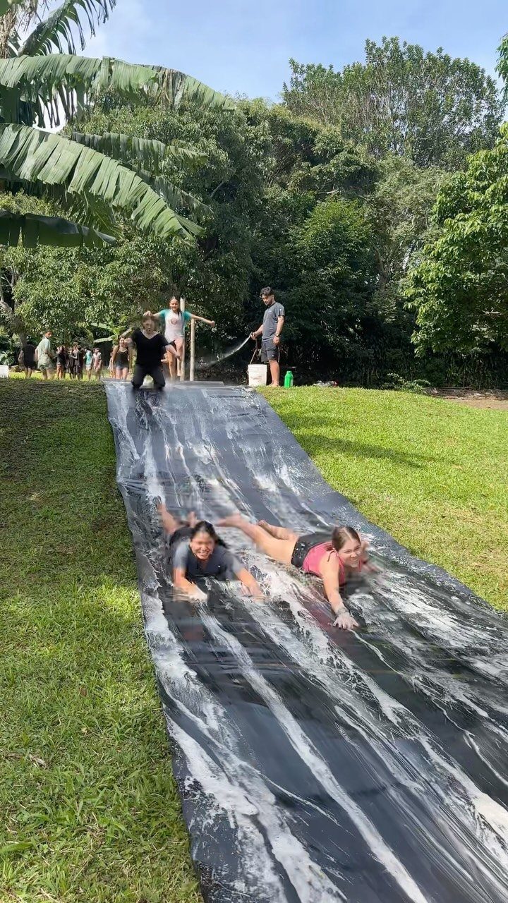 Two women sliding down a long black plastic slip-and-slide on a grassy lawn