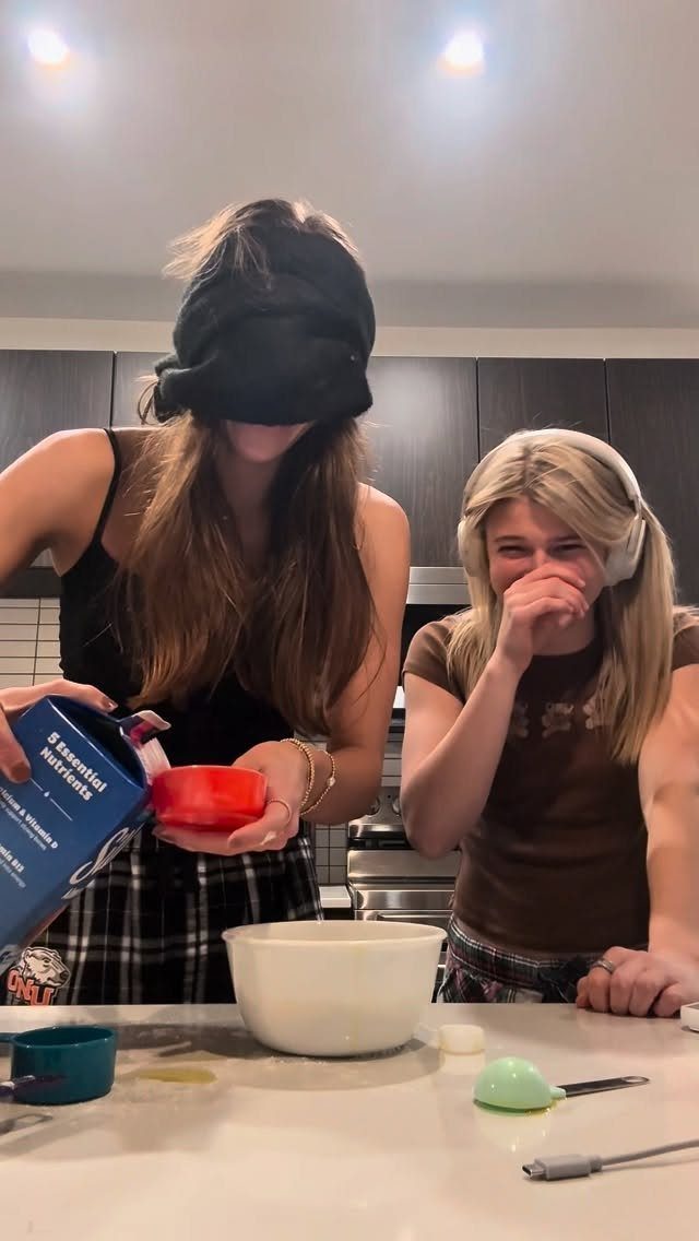 Blindfolded woman pouring ingredients into a bowl while her friend laughs in kitchen