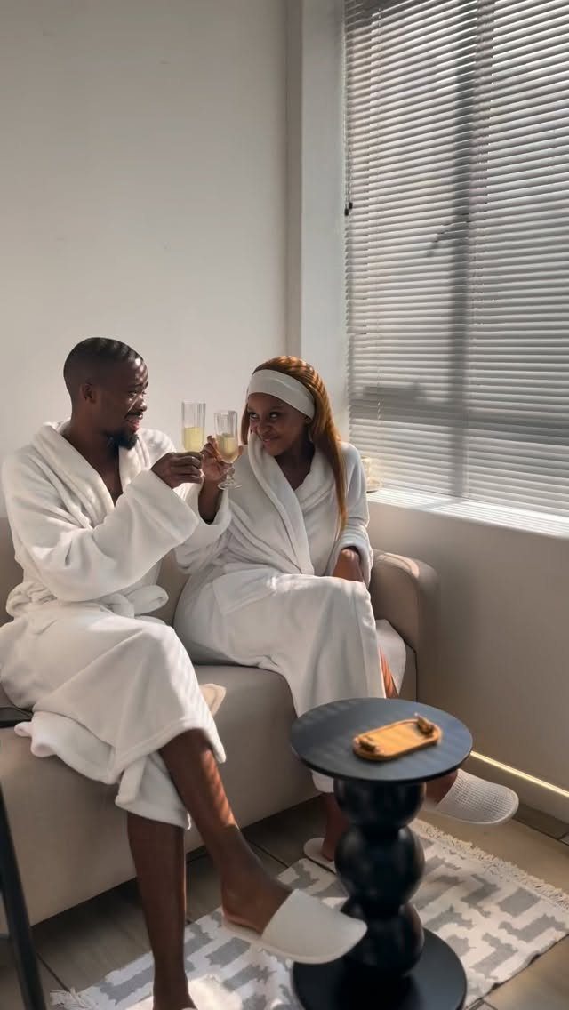Couple in white bathrobes clinking champagne glasses while sitting on a sofa.