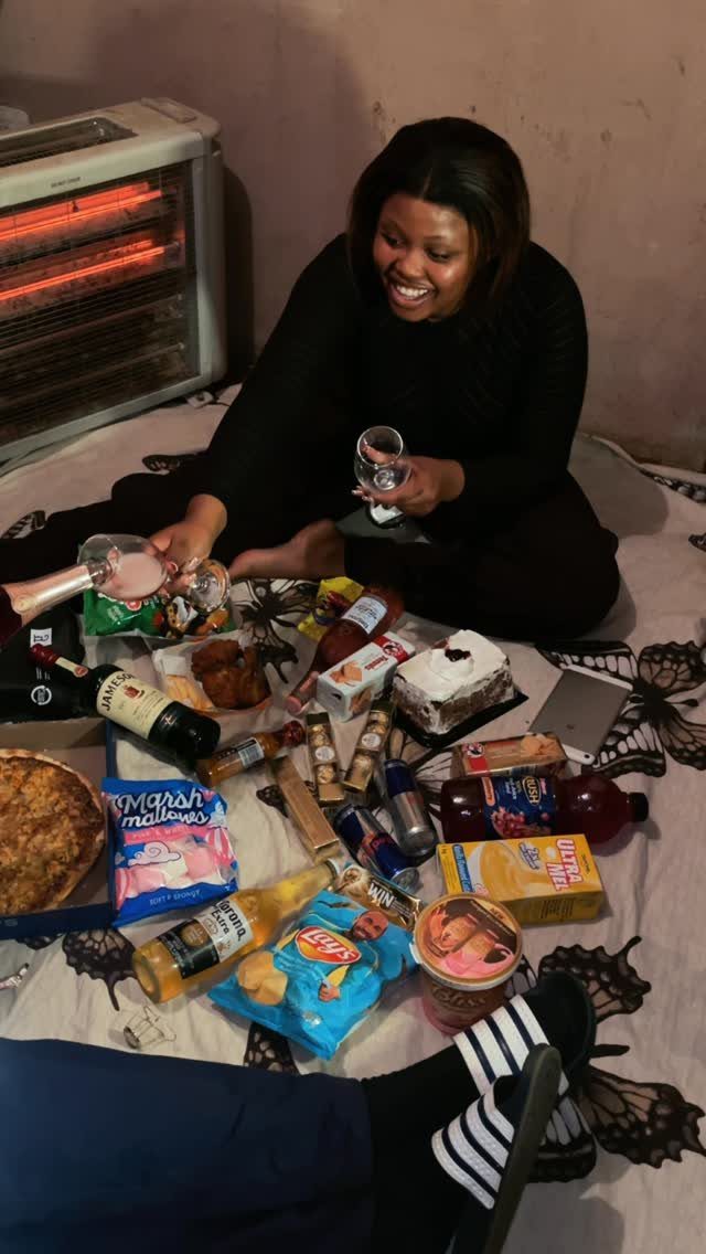 Woman smiling while sitting on a floor blanket surrounded by snacks and drinks.