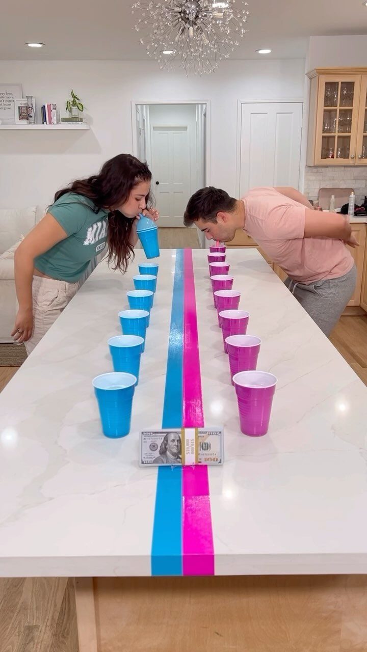 Man and woman blowing plastic cups across a white table during a party game.