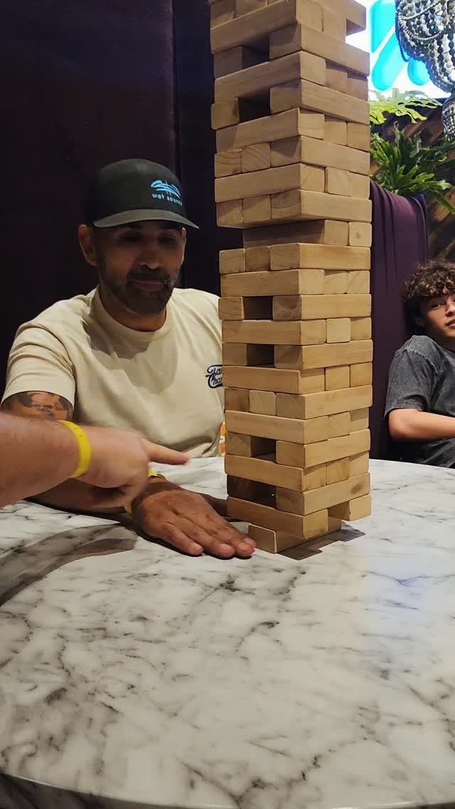 Tall wooden block tower on a marble table with people watching closely.