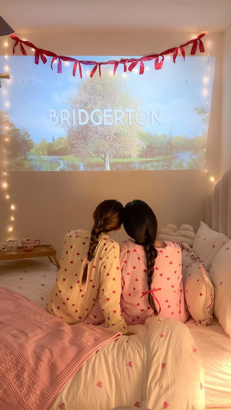 Two women in heart pajamas watching a movie projection in a decorated bedroom.