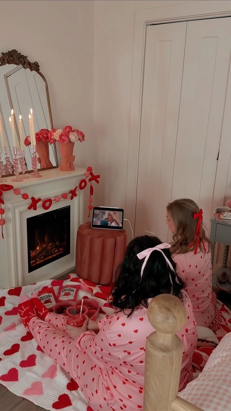 Two people in pink heart pajamas sitting on a heart blanket near a fireplace.