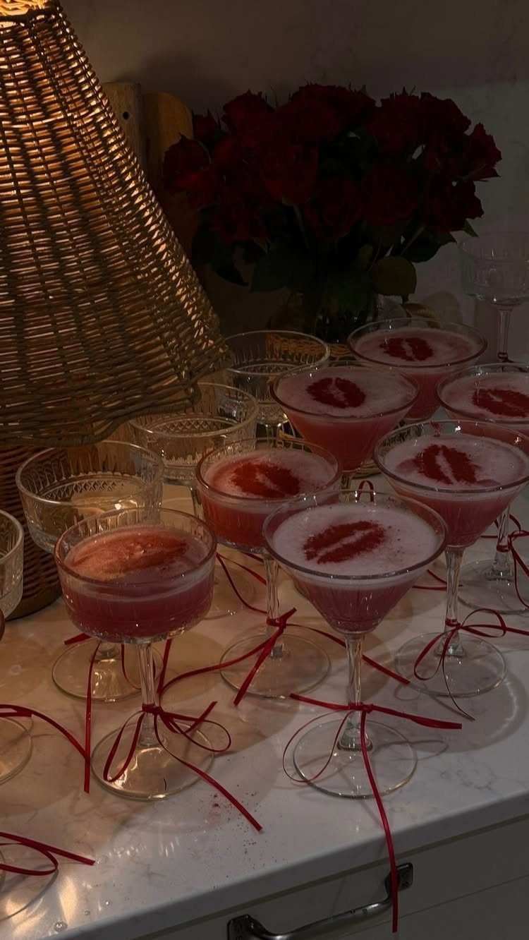 Pink cocktails with red lip garnishes and ribbons next to red roses.