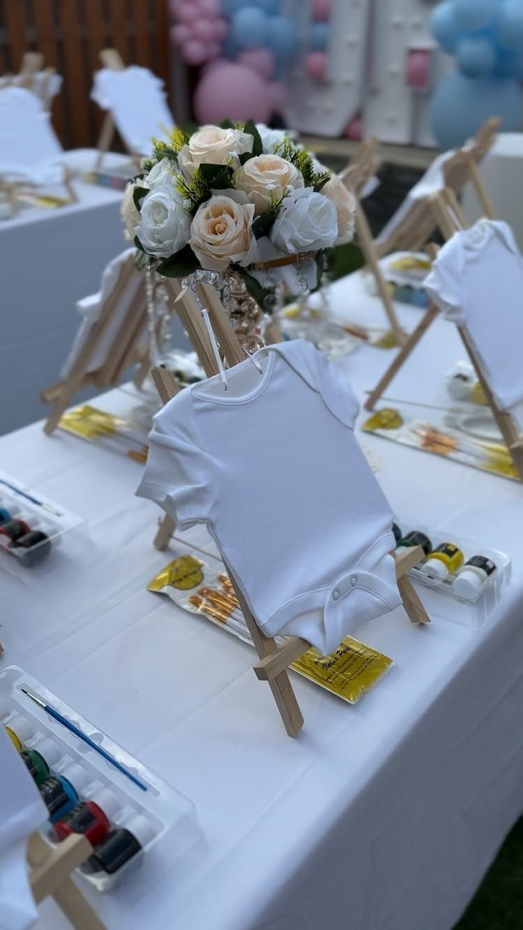 White baby onesies on wooden easels with paint pots and floral centerpiece.