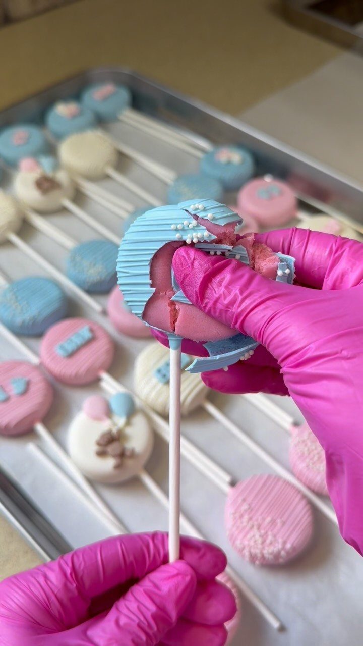 Broken blue cake pop revealing pink filling held by pink gloved hands.
