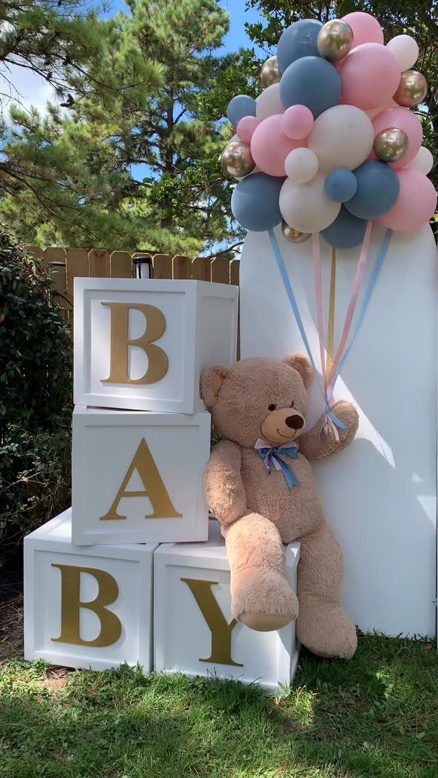 White BABY blocks, a brown teddy bear, and pink and blue balloons outdoors.