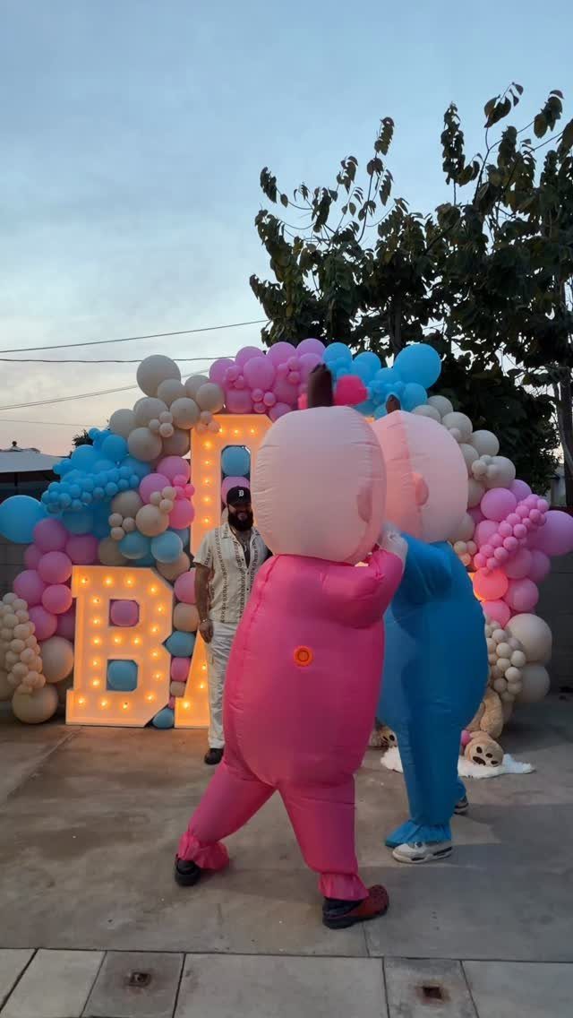 Inflatable pink and blue costumes posing before a baby-themed balloon arch and lights.