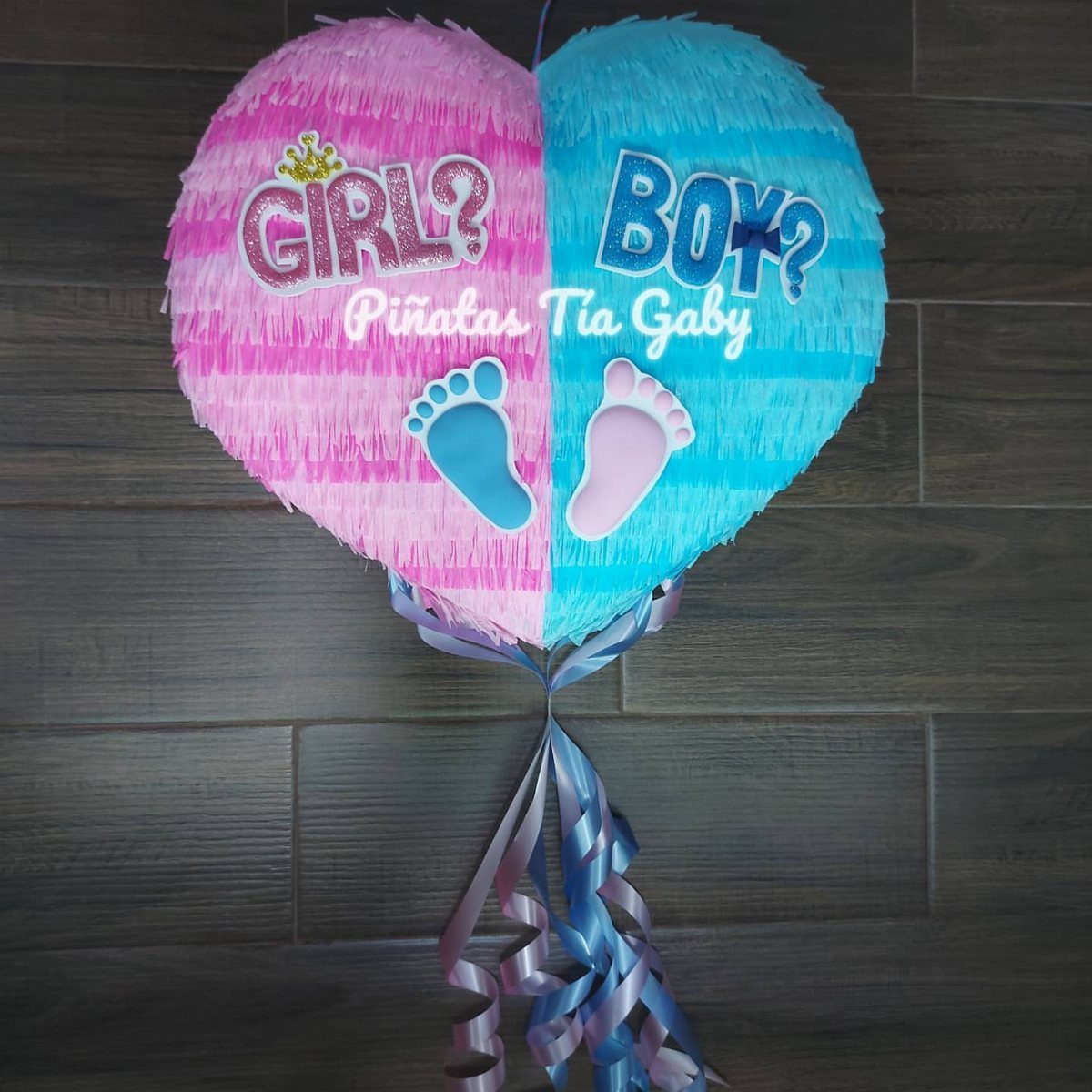 Heart-shaped pink and blue gender reveal piñata with baby footprints on wood wall