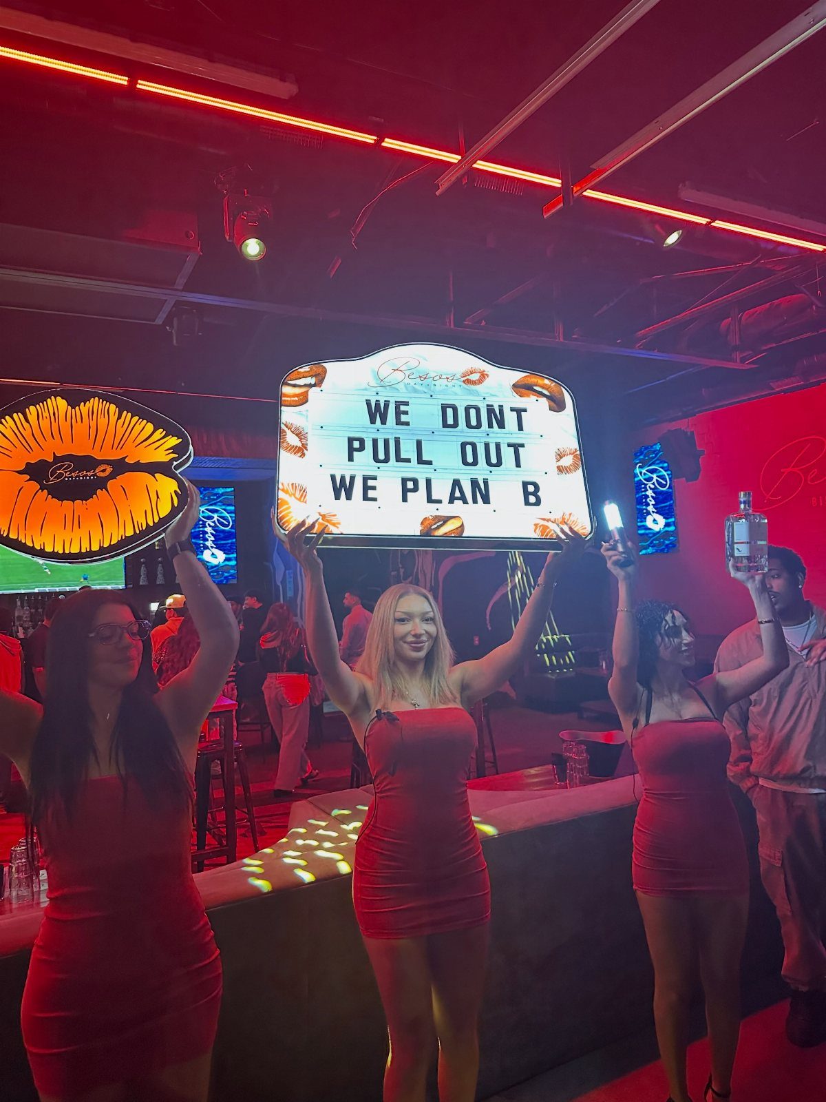 Three women wearing red mini dresses posing in a red-lit nightclub setting.