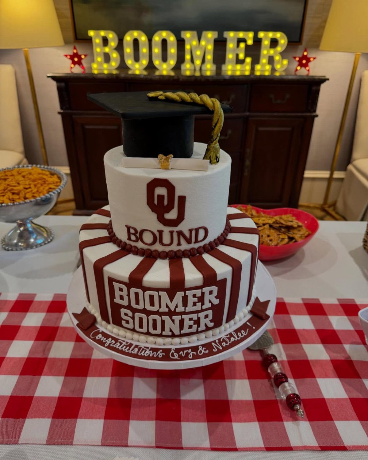 Graduation cake with a black cap topper on a red checkered tablecloth.