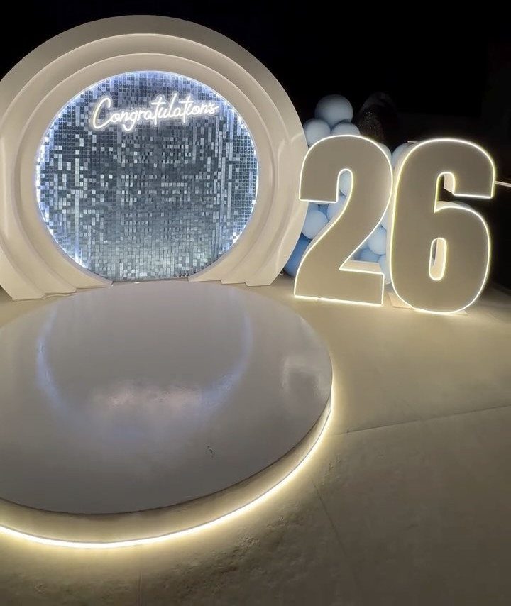Celebratory event setup with shimmering backdrop, neon congratulations sign, and glowing number 26.