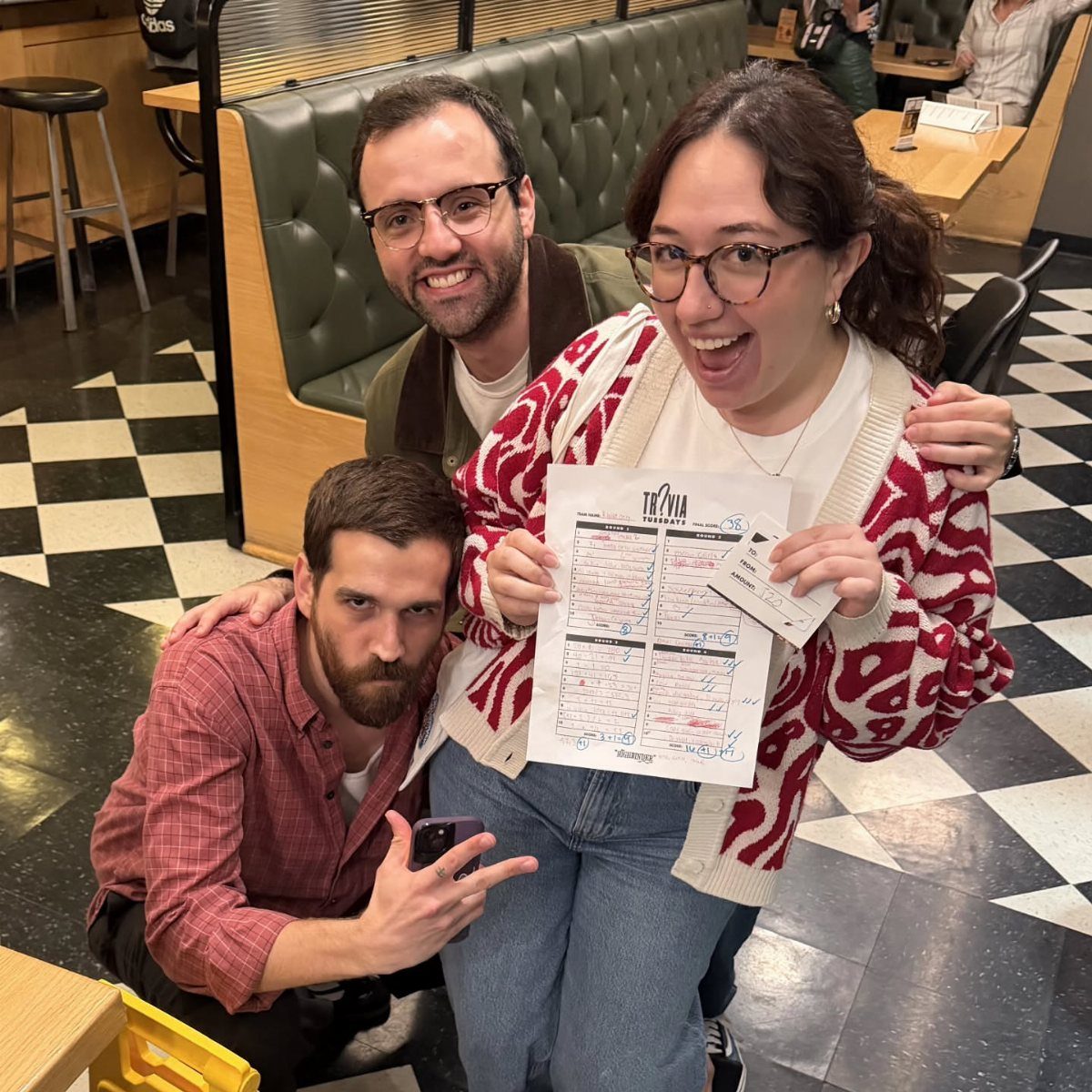 Three friends posing in a diner, holding a trivia night answer sheet.