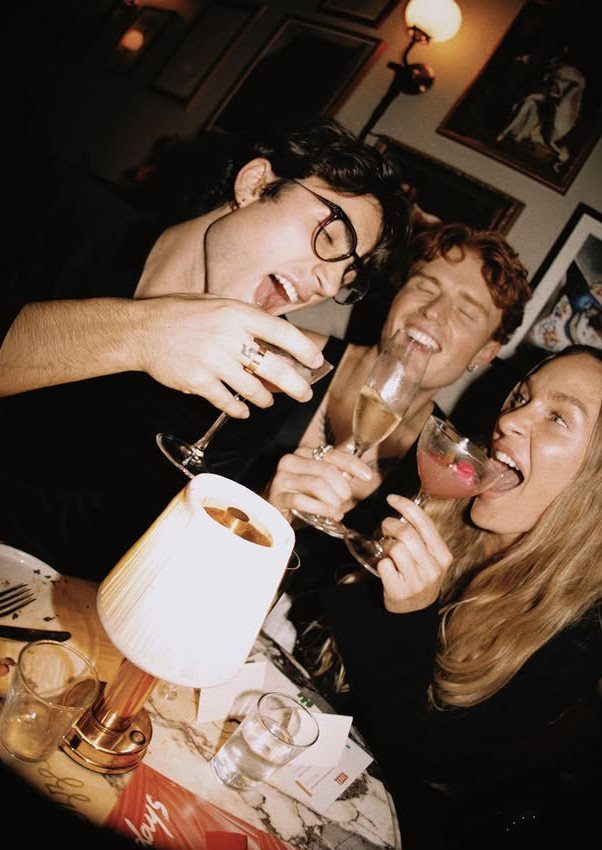 Three friends laughing and drinking cocktails at a table in a cozy room.