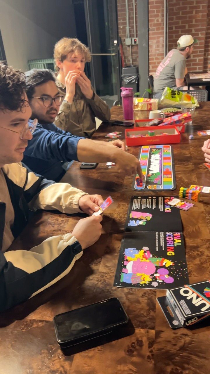 Young men playing a colorful board game around a wooden table indoors.