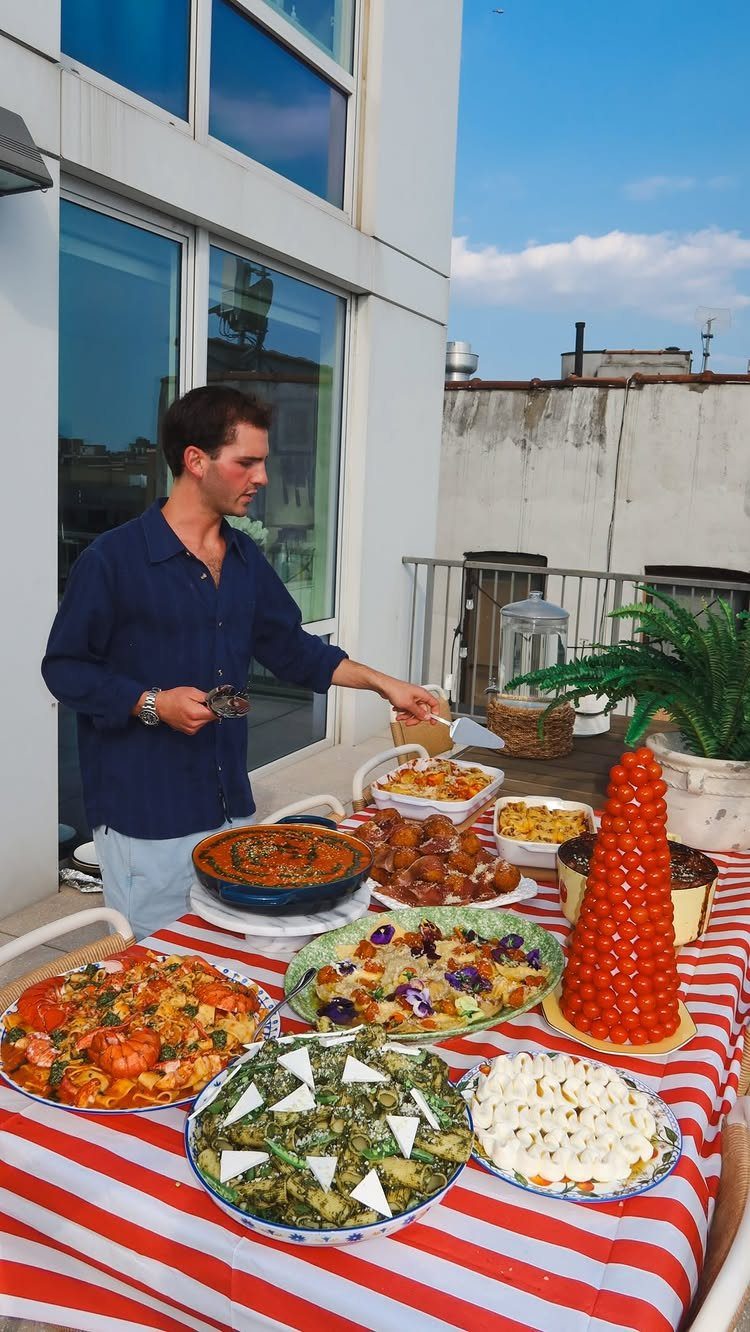 Man standing by a table of diverse homemade dishes on a sunny rooftop terrace.