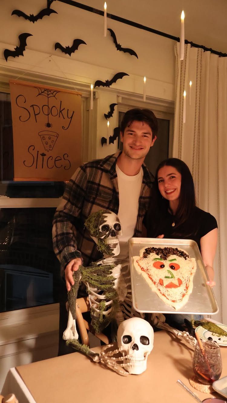 Couple posing with a face-shaped pizza and skeleton for a spooky themed dinner.