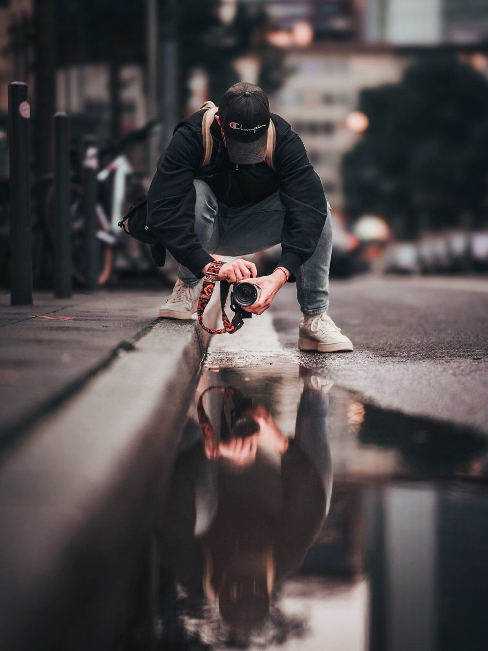 Person crouching on a city street taking a photo of a puddle reflection.