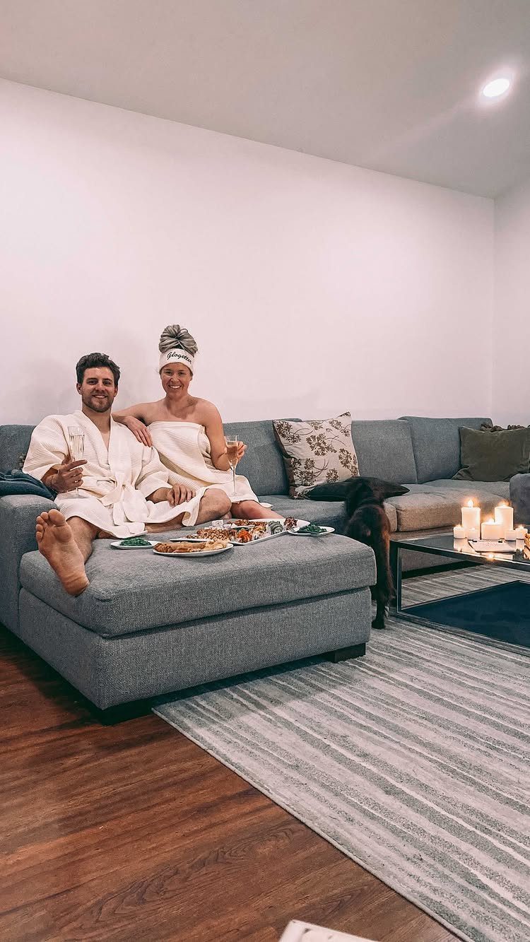 Couple in white bathrobes relaxing on a grey sofa with candles and snacks.