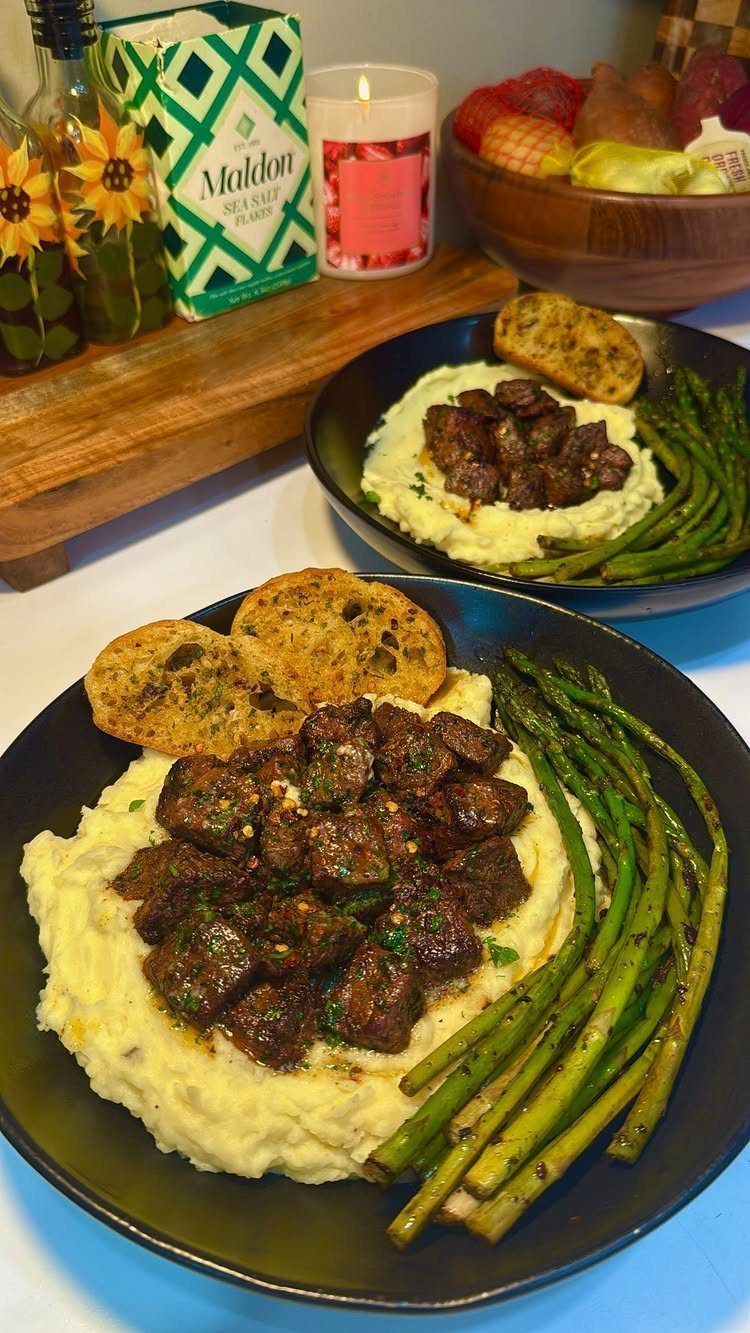 Steak tips with mashed potatoes and asparagus served on two black plates.