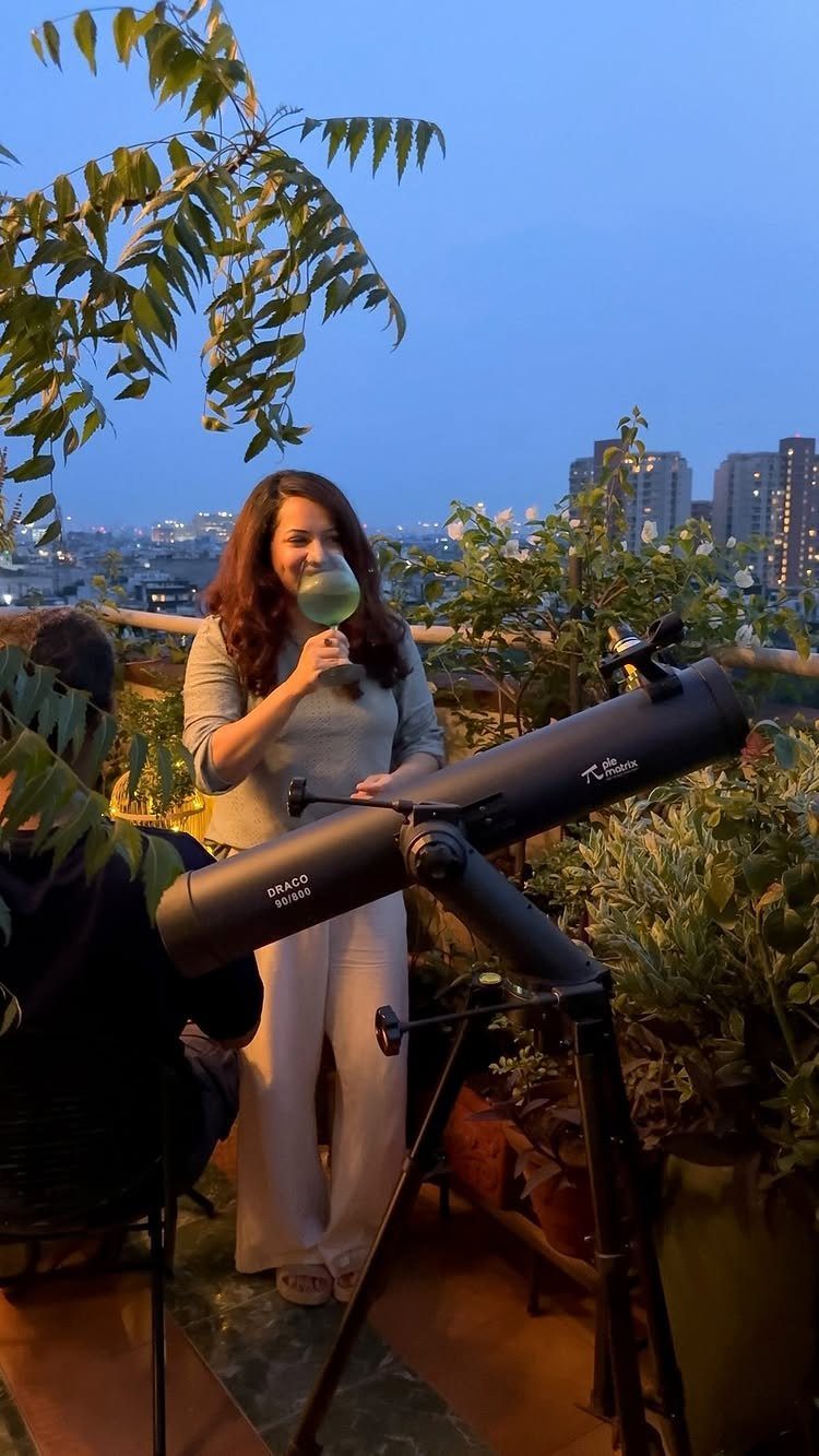 Woman holding a wine glass standing next to a telescope on a rooftop balcony.