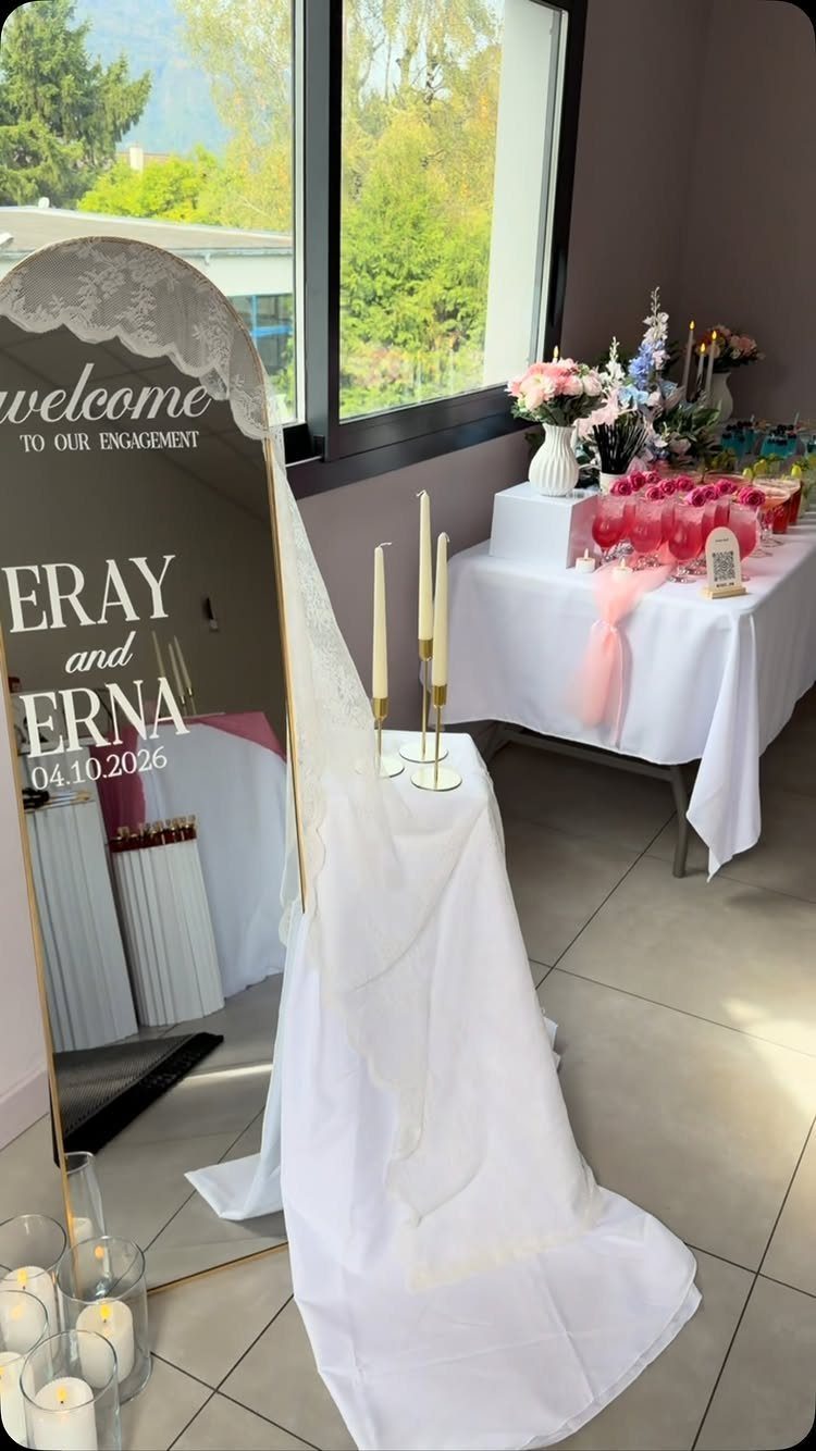 Engagement party decor with a lace-trimmed mirror, white drapery, and pink cocktail table.
