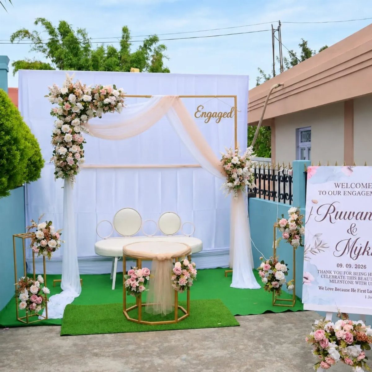 Outdoor engagement party setup with white floral arches, peach drapes, and gold furniture.
