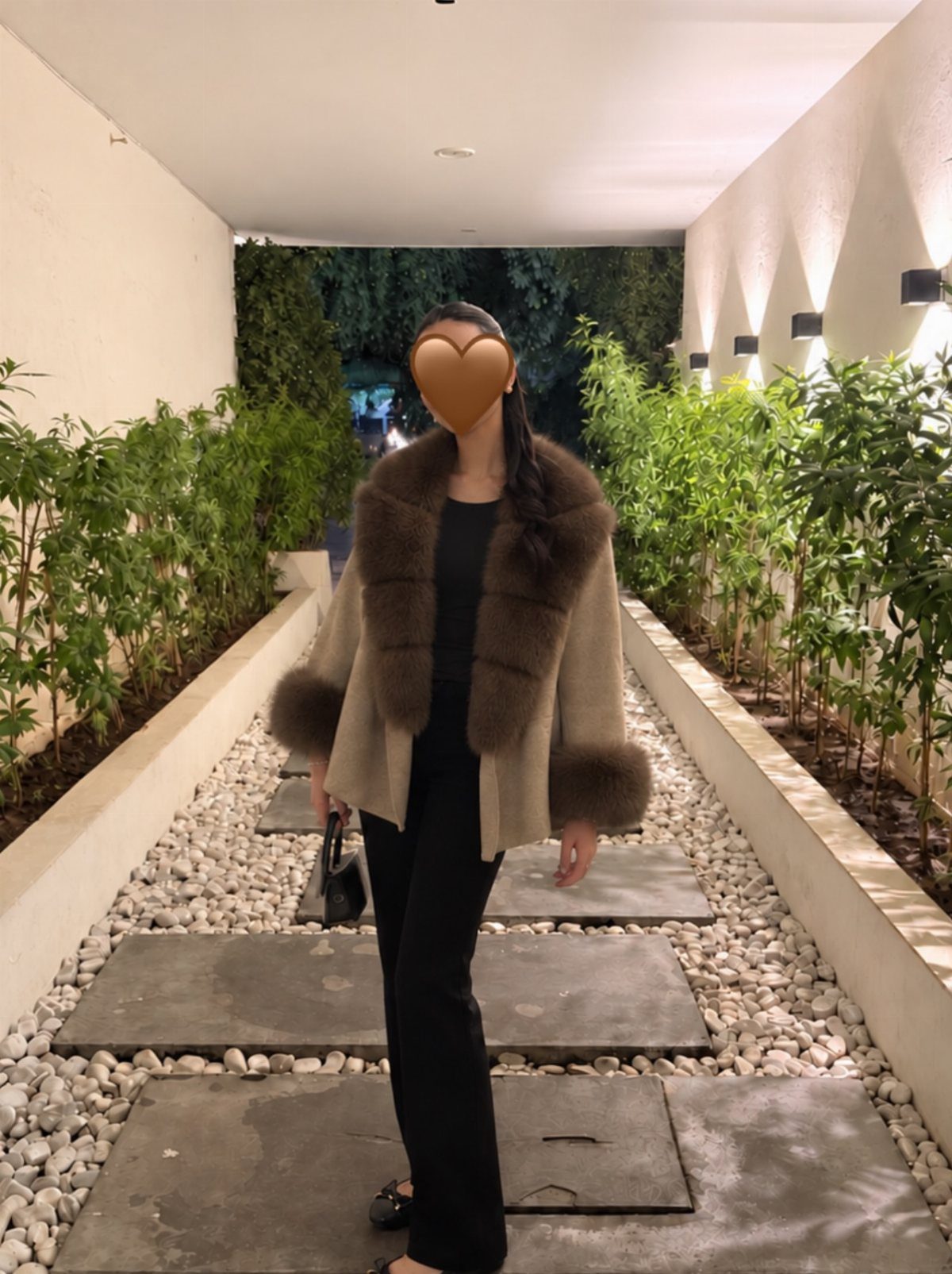 Person wearing a beige faux fur trimmed jacket and black straight-leg trousers outdoors.