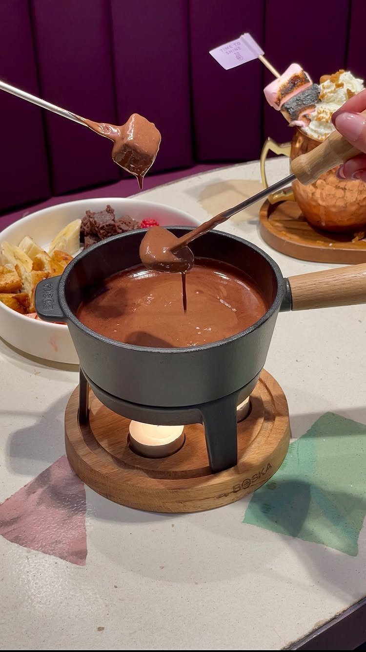 Chocolate fondue pot with dipping skewers and fruit on a white table.