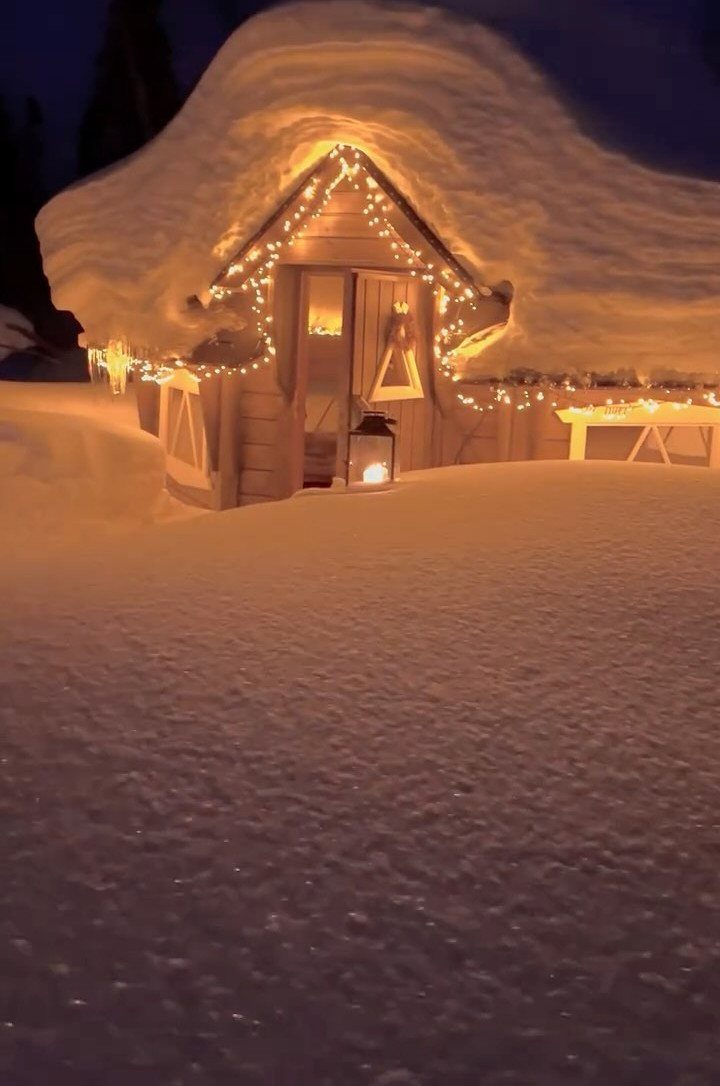 Snow-covered wooden cabin at night illuminated by warm yellow string lights and lanterns.
