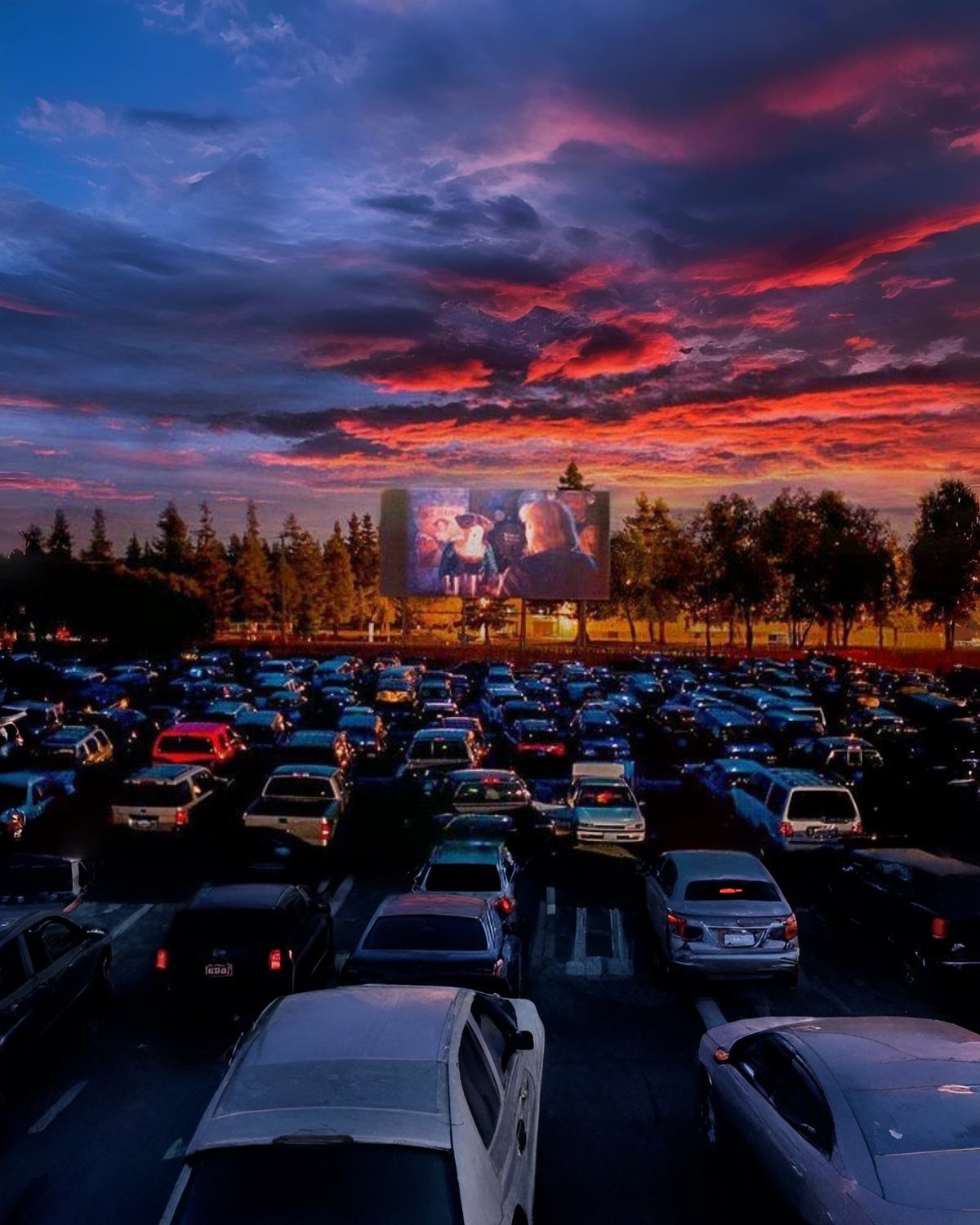 Outdoor movie screen at a drive-in theater with cars under a sunset sky.