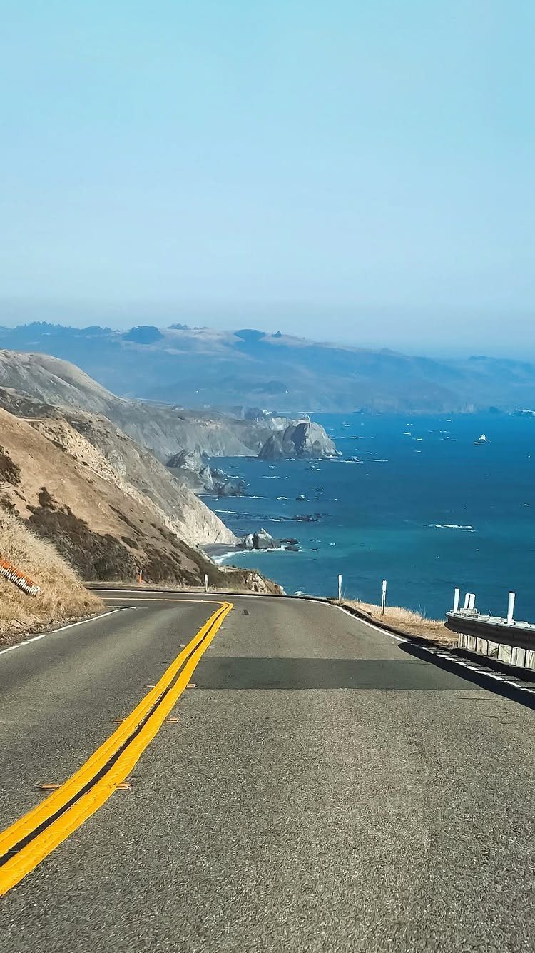Winding coastal road with yellow lines overlooking a blue ocean and mountains.