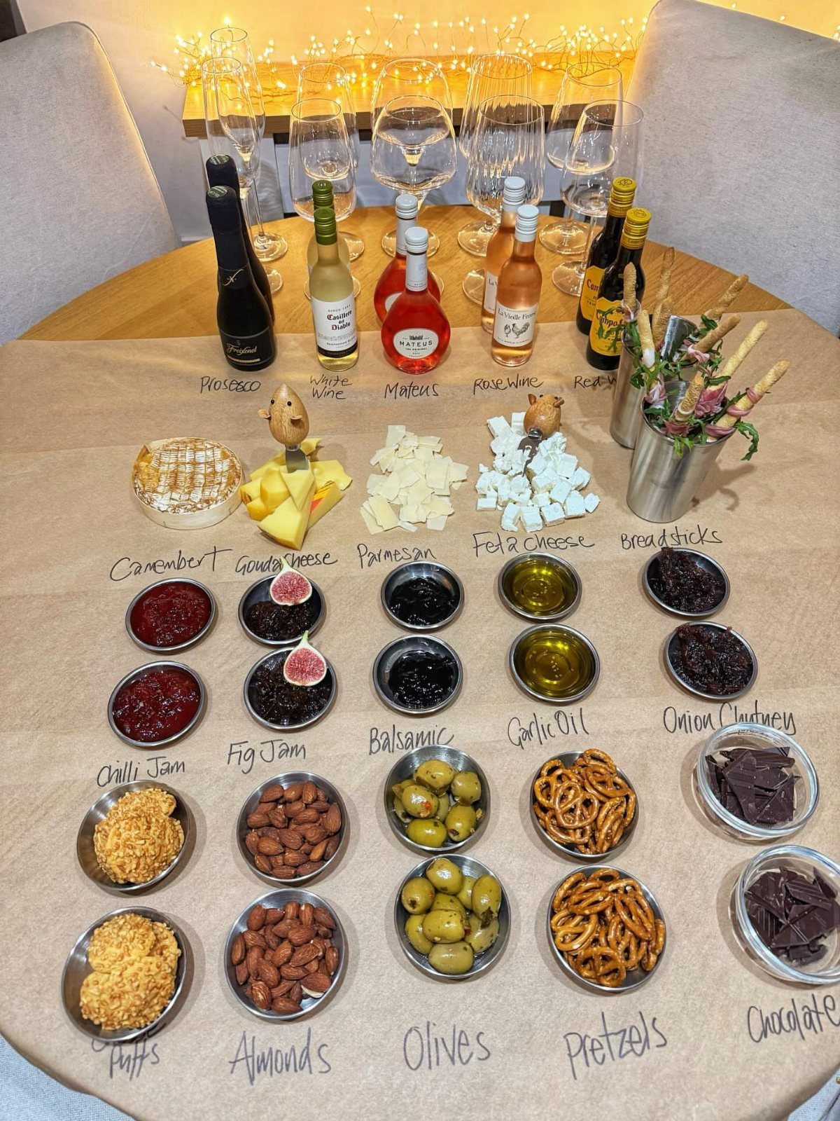 Wine and charcuterie spread with labeled bowls of snacks on a brown paper table.
