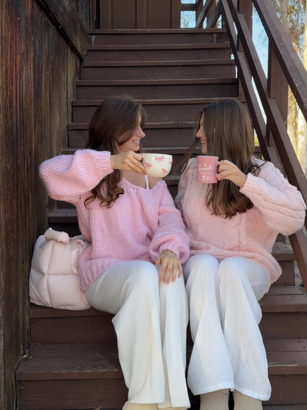 Two women in matching oversized pink sweaters and white pants sitting on wooden stairs.