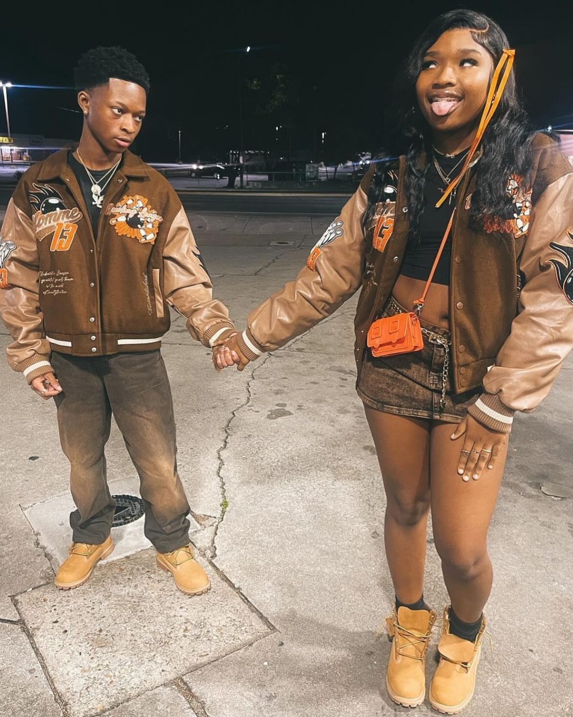 Young couple holding hands wearing matching brown varsity jackets and tan boots outdoors.