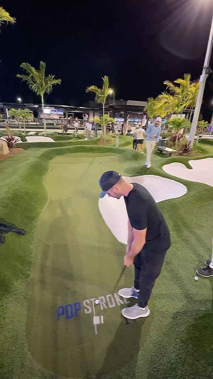 Man in black clothing putting a golf ball on a night mini golf course.