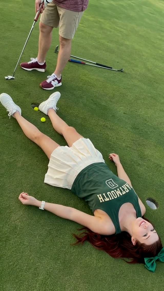 Woman lying on green turf next to a man holding a golf putter.