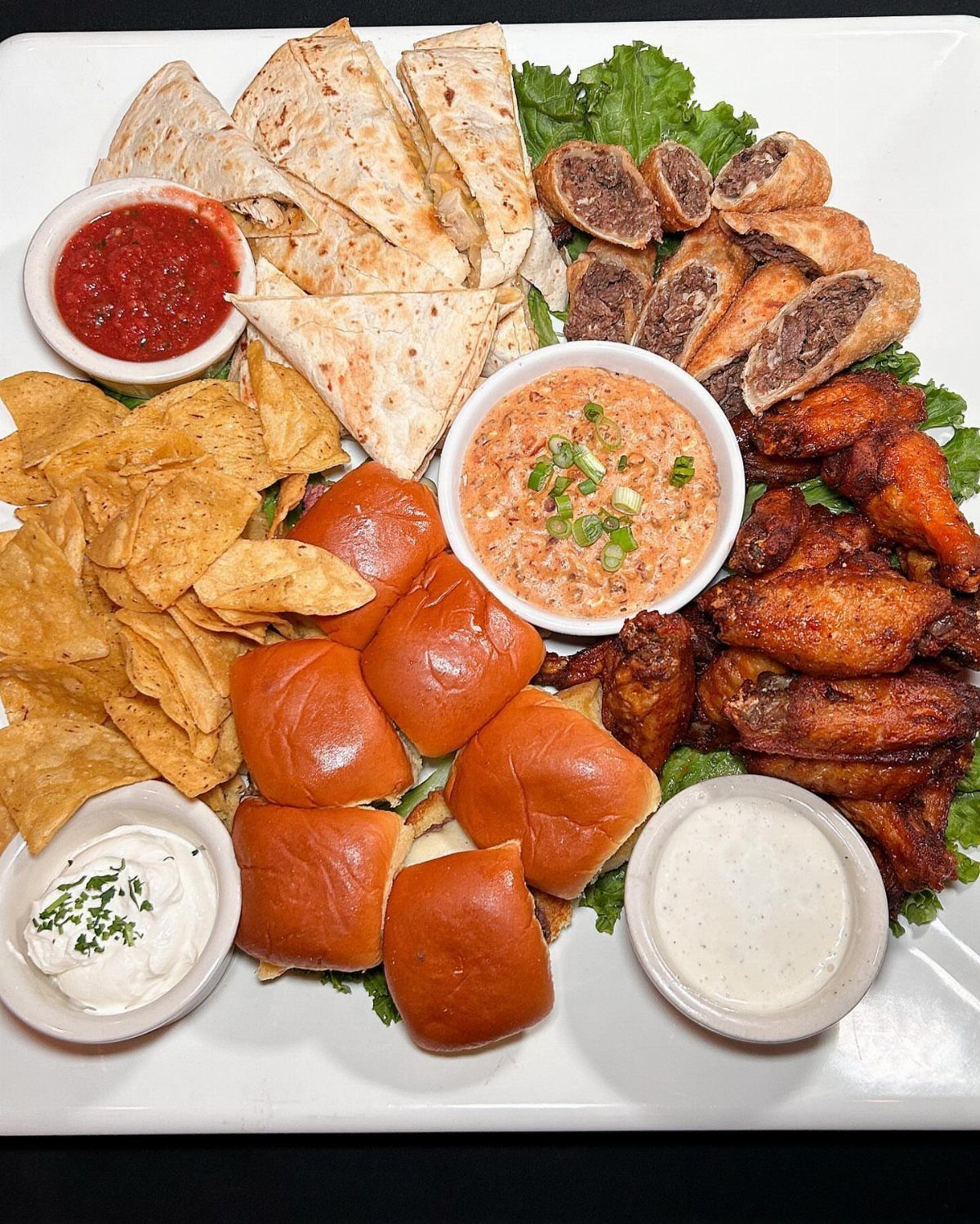 Appetizer platter with wings, sliders, chips, and dips on a white plate.