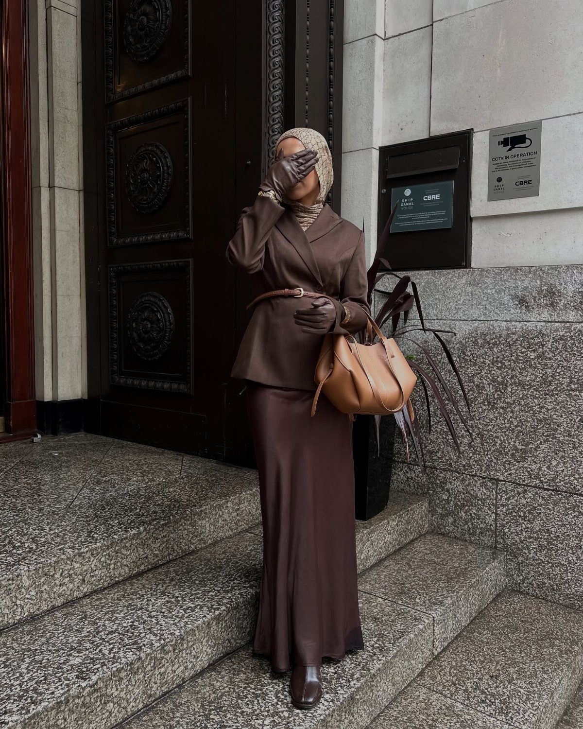Woman in brown blazer and satin skirt wearing a beige hijab on stone steps