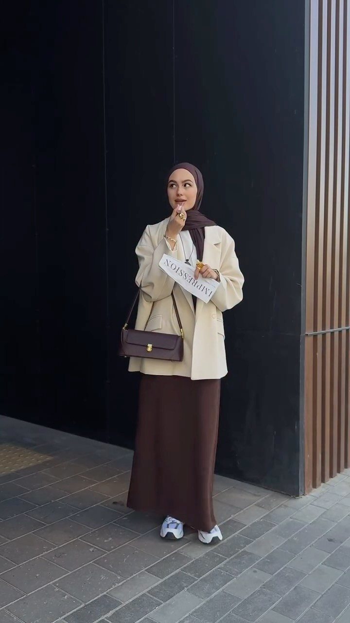 Woman in cream blazer and brown maxi skirt posing against a dark wall.