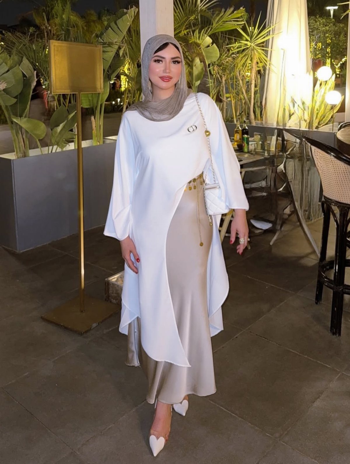 Woman in champagne satin maxi dress, white cover-up, and grey hijab standing outdoors.
